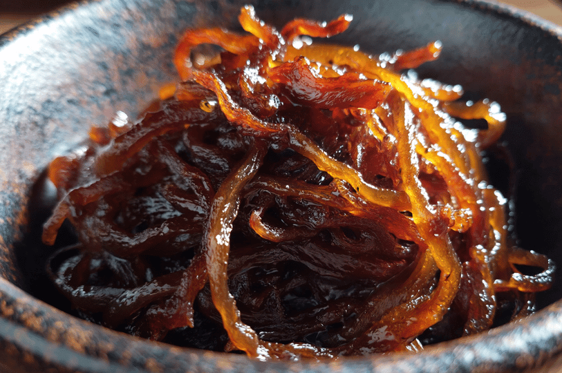 An extreme close-up of dark, balsamic-glazed caramelized onions in a ceramic bowl, highlighting the sticky, translucent, and concentrated nature of long-cooked onions.