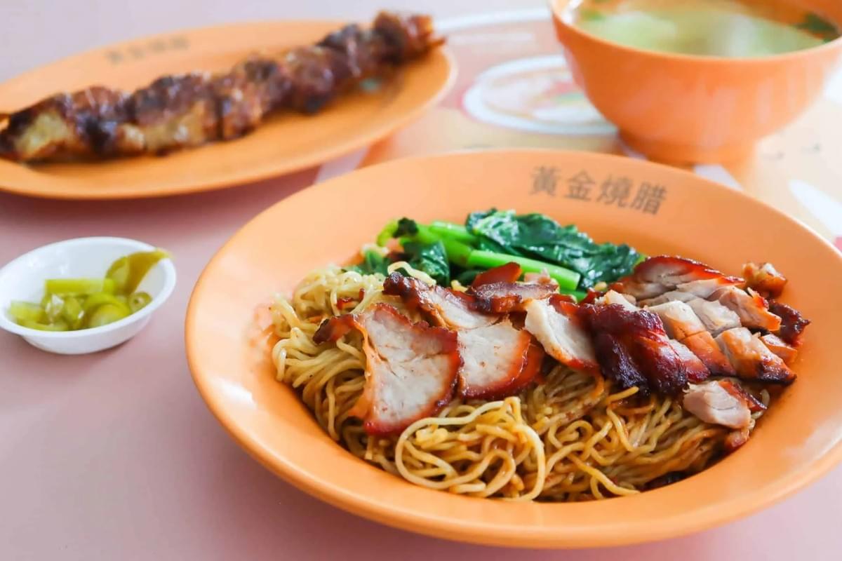 A bowl of thin egg noodles is topped with sliced char siew pork and bright green vegetables. In the background, a plate of roasted meat and a small bowl of soup complete the meal set.