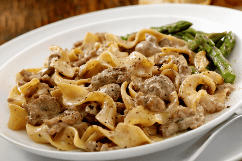 A warm, close-up angled shot of hearty Beef Stroganoff mixed with twisted egg noodles, seasoned with cracked black pepper, and served alongside bright green steamed asparagus.