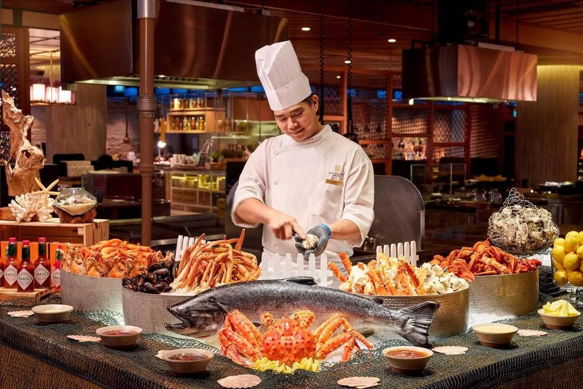 A professional chef in a white uniform and tall hat stands behind a lavish seafood buffet counter, meticulously shucking an oyster. The display in front of him features an abundant arrangement on ice, including a whole large salmon, king crabs, lobsters, shrimp, and various shellfish, all set within a warmly lit, modern restaurant environment.