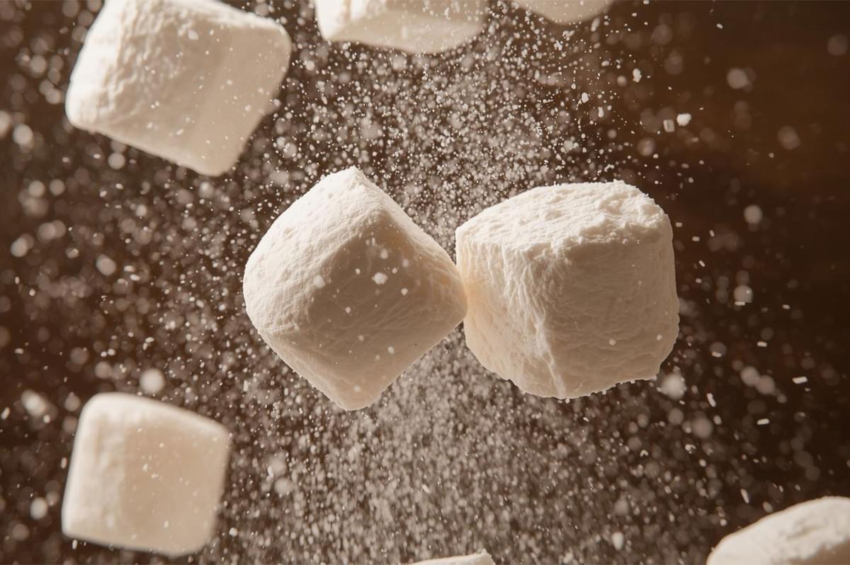 Dynamic shot of two square white marshmallows suspended in mid-air, with a cloud of powdered sugar falling around them, suggesting their light, airy quality.