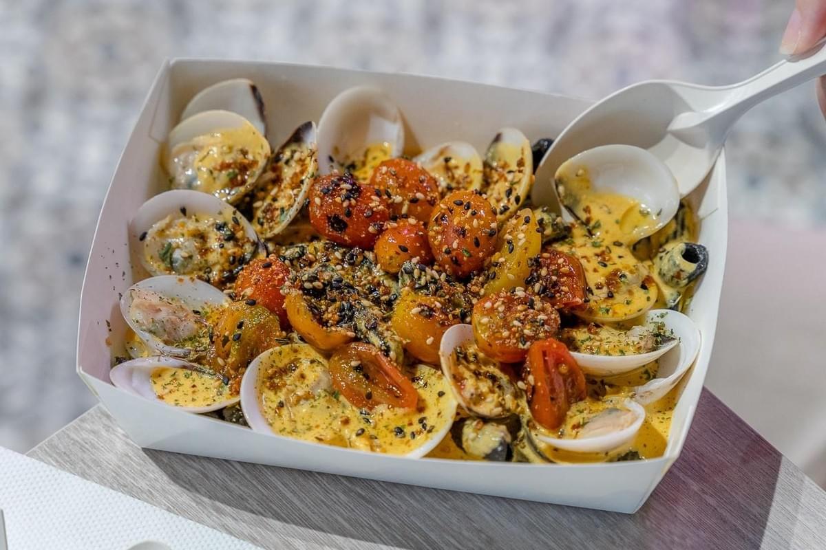 A white takeaway box holds a creamy pasta dish loaded with open-shell clams and halved cherry tomatoes. The meal is garnished with sesame seeds and herbs, with a white plastic spoon scooping up a portion from the container.