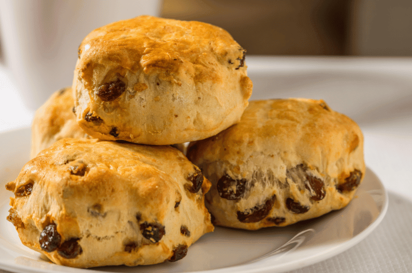 A close-up stack of fruit scones on a white plate, showing golden crusts and a generous amount of dark raisins baked into the dough.