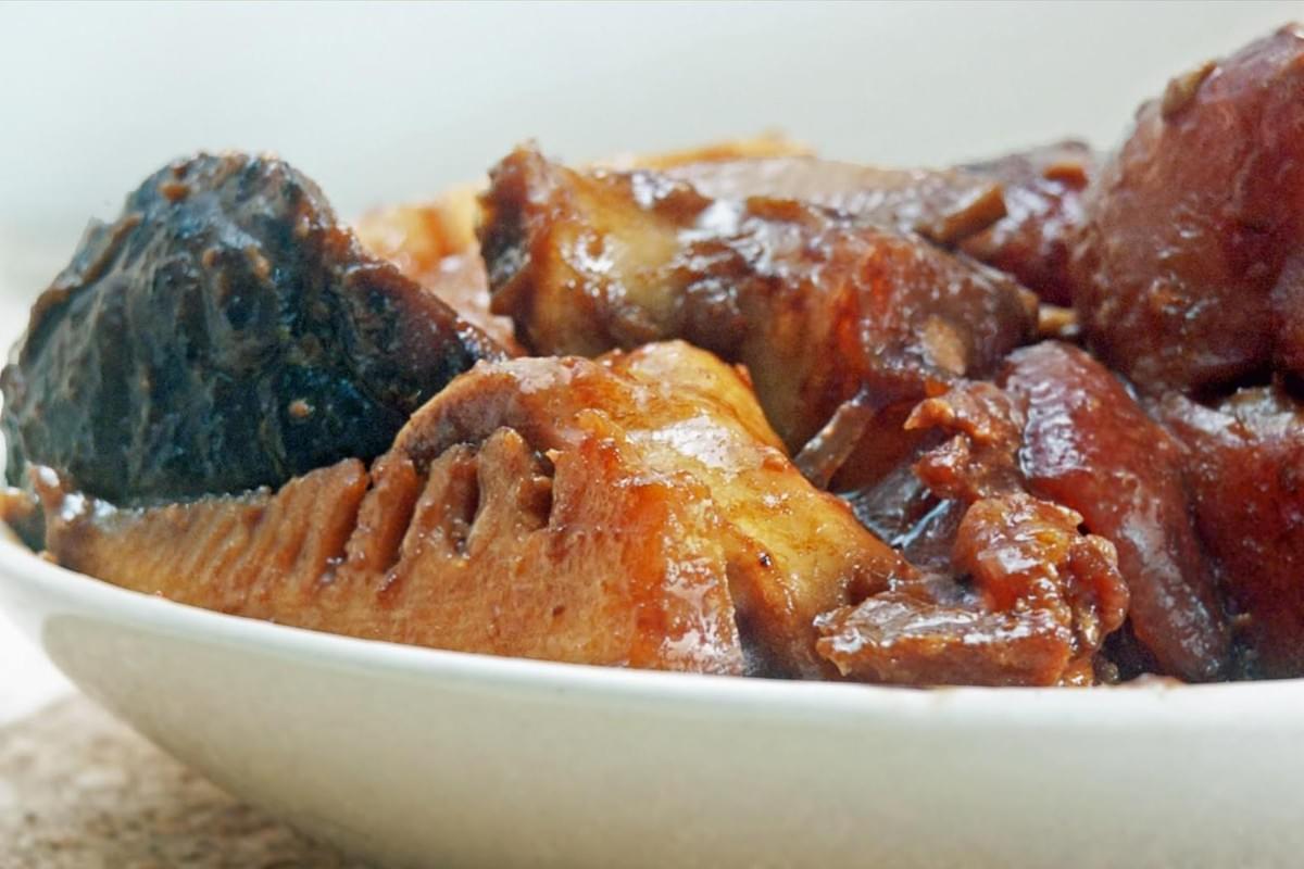  Gemini said A close-up shot features a white bowl filled with Babi Pongteh, a traditional Peranakan braised pork dish known for its rich, savory gravy. The image highlights tender chunks of pork belly and bamboo shoots coated in a dark soy and fermented bean sauce, accompanied by whole shiitake mushrooms.