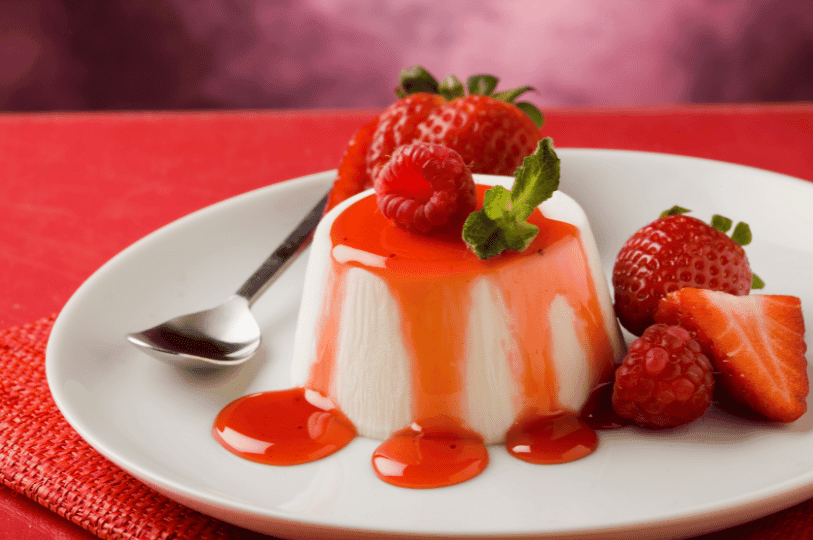 A classic unmolded vanilla Panna Cotta on a white plate, drizzled with vibrant red strawberry coulis and garnished with fresh strawberries and a mint leaf.