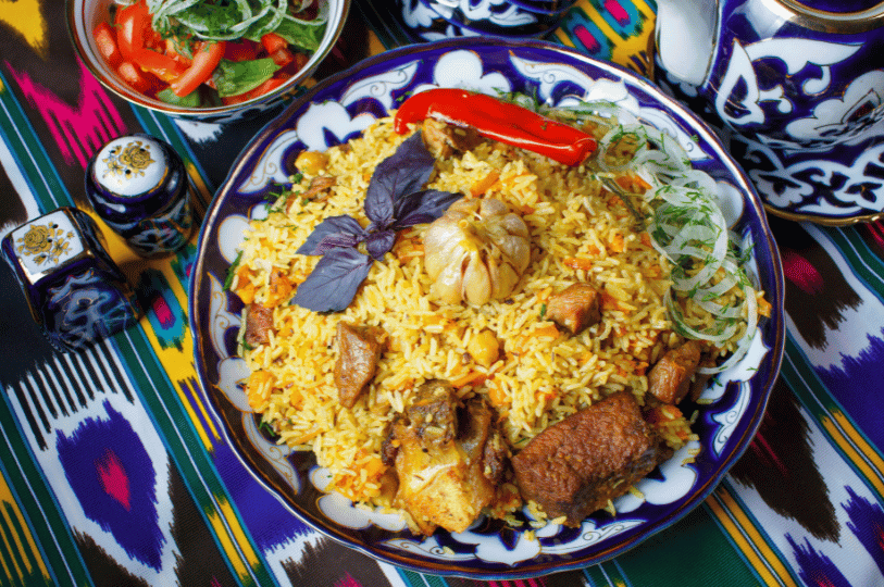 Authentic Uzbek plov served on a traditional blue and white lagan plate, prominently featuring large chunks of meat, a whole roasted garlic head, red chili, and fresh purple basil, resting on a vibrant ikat fabric.