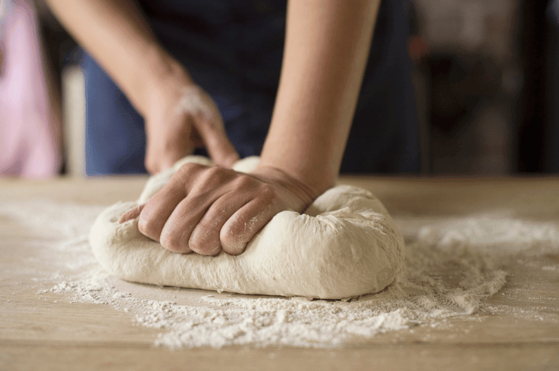 A baker using their palms to press and stretch smooth bread dough on a wooden table, a key technique for strengthening the gluten network.
