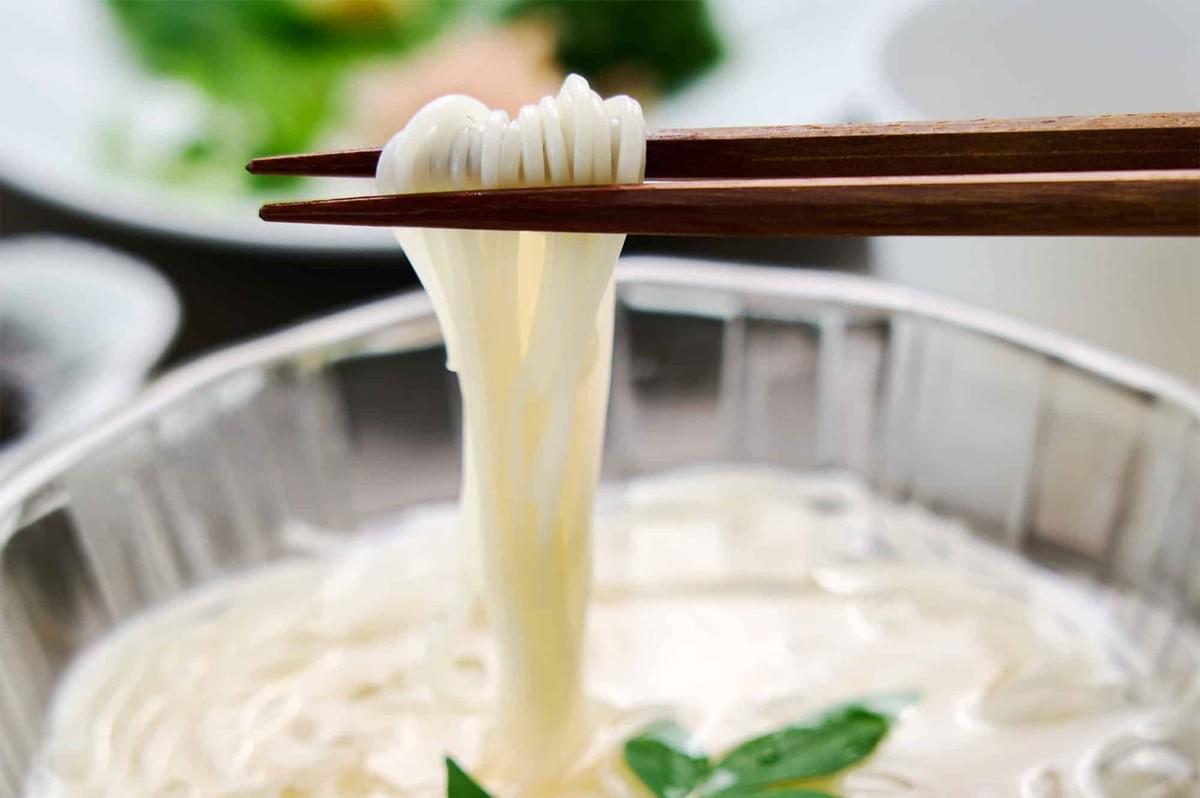 Wooden chopsticks lifting a portion of thin, glossy white noodles from a bowl, showing the smooth texture of the pasta.