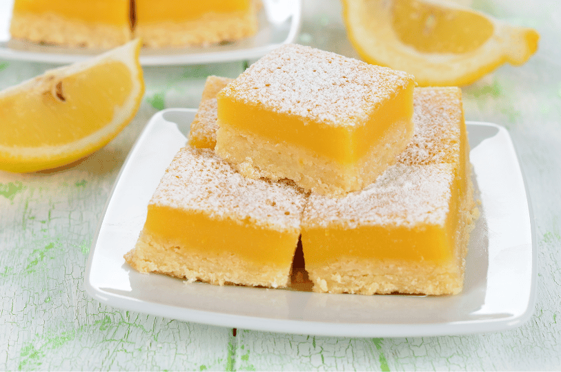 A close-up stack of three thick, square lemon bars on a white plate. Each bar features a bright yellow lemon curd layer atop a buttery shortbread crust, heavily dusted with powdered sugar and accented by fresh lemon wedges.