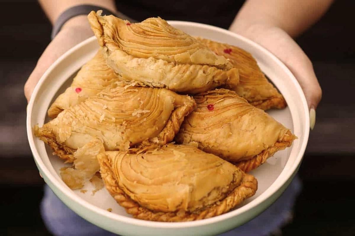 Plate of golden-brown pastries with flaky, layered crusts, held by hands. The pastries have a rustic, homemade appearance and are garnished with small red bits.