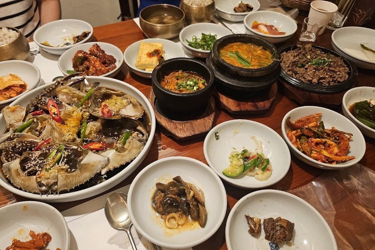 The image displays a wooden dining table covered in a rich variety of traditional Korean dishes served in numerous bowls. Among the spread are raw marinated crabs garnished with peppers, hot stews bubbling in black earthenware pots, marinated beef bulgogi, and an assortment of colorful side dishes (banchan).