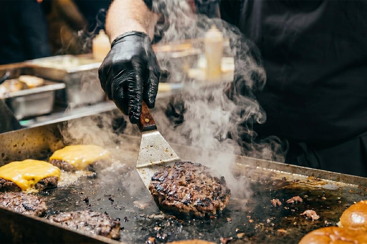 Chef flipping a smash burger patty on a hot flat top grill with melting cheese and steam.