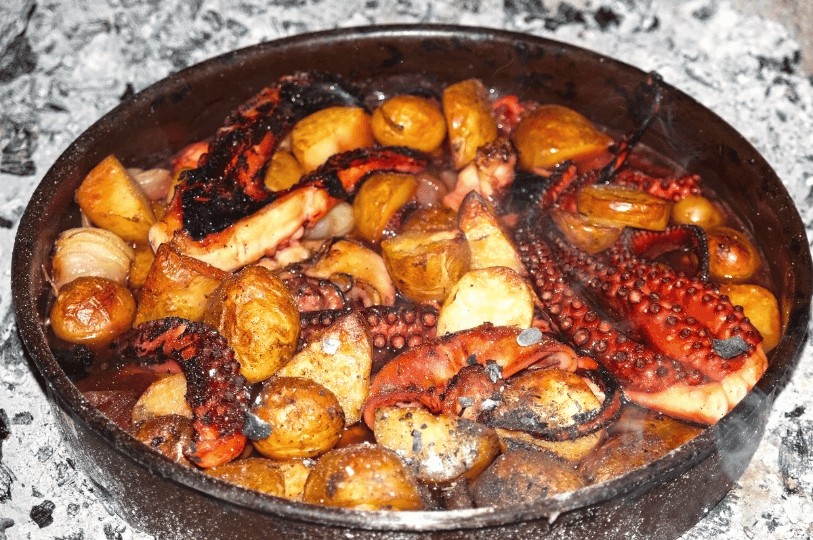 A traditional dark Peka pan resting directly in hot white wood embers, filled with slow-roasted octopus tentacles and halved potatoes.