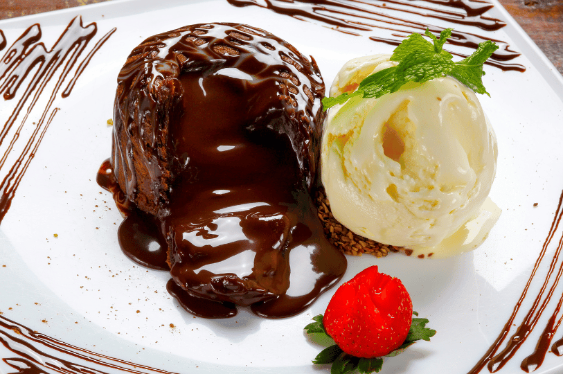 A decadent molten lava cake being served with a large scoop of vanilla bean ice cream. Thick chocolate sauce flows from the top and center of the cake, surrounded by artistic chocolate drizzles and a whole fresh strawberry.