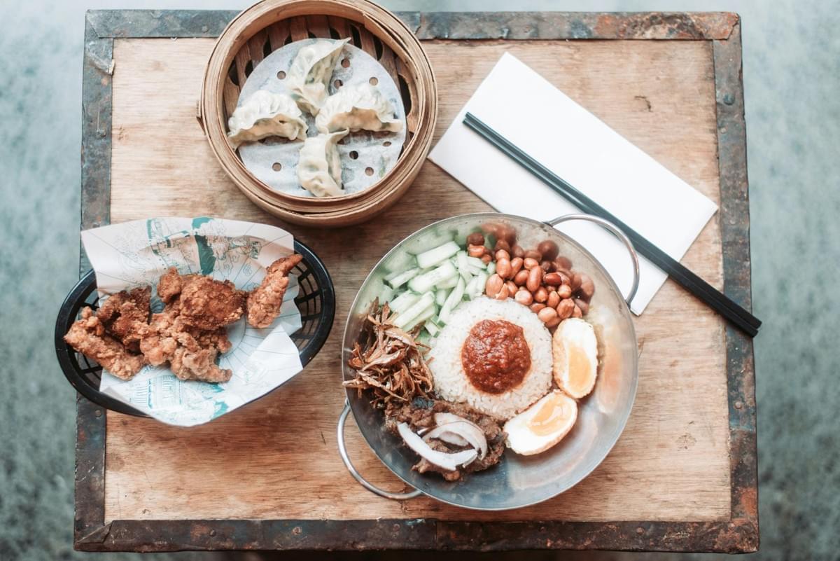 An overhead view shows a rustic wooden table set with three Asian dishes, including steamed dumplings in a bamboo basket, crisp fried chicken, and nasi lemak topped with sambal sauce. Chopsticks rest on a white napkin next to the metal wok containing rice, eggs, peanuts, anchovies, and sliced cucumbers.