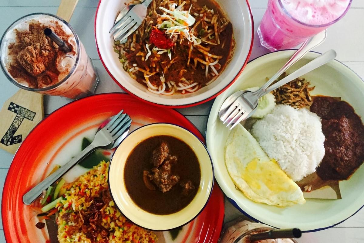 A vibrant spread of Malaysian dishes is laid out on a table, including Nasi Lemak with a fried egg, flavorful noodles, and a rich meat curry. The meal is accompanied by classic local beverages like a tall glass of iced Milo topped with powder and a bright pink Sirap Bandung.