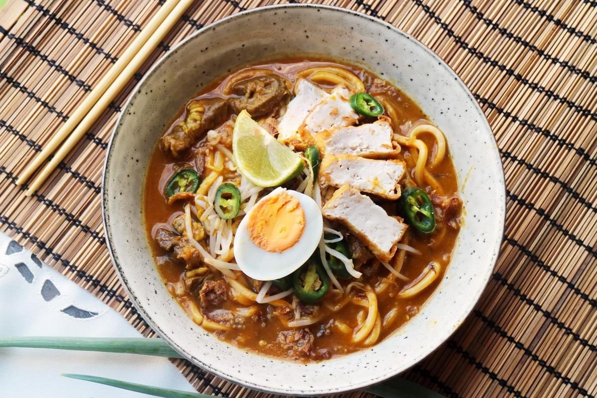 A ceramic bowl filled with Mee Rebus noodle soup rests on a woven bamboo mat next to a pair of wooden chopsticks. The dish features yellow noodles in a thick orange gravy, topped with sliced hard-boiled egg, lime, fried tofu, bean sprouts, and green chilies.