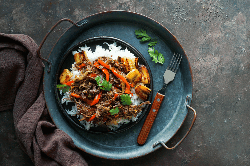 An overhead view of a modern plating of Ropa Vieja served on a bed of white rice, garnished with vibrant red bell pepper strips, fresh cilantro, and golden fried plantains on a dark metal tray.