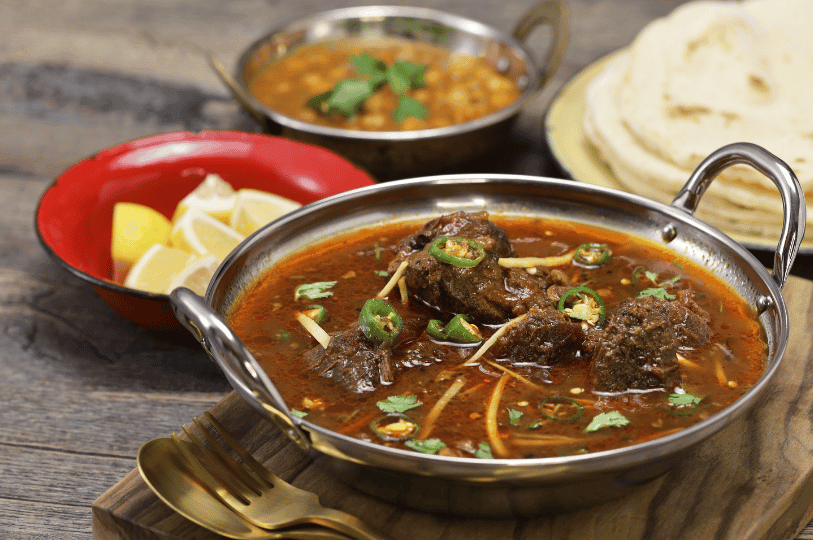 A full table spread featuring a metal bowl of Nihari, a side of lemon wedges in a red dish, a small bowl of lentils, and golden-brown naan.