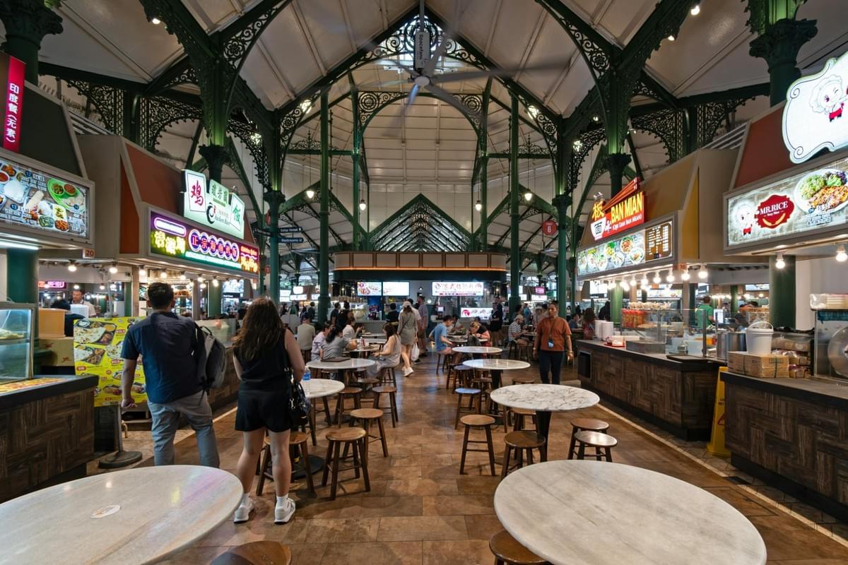 A wide view inside Lau Pa Sat hawker center in Singapore, featuring green ornate cast-iron columns and a high vaulted ceiling. Diners walk past circular wooden tables while various illuminated food stalls line both sides of the hall.