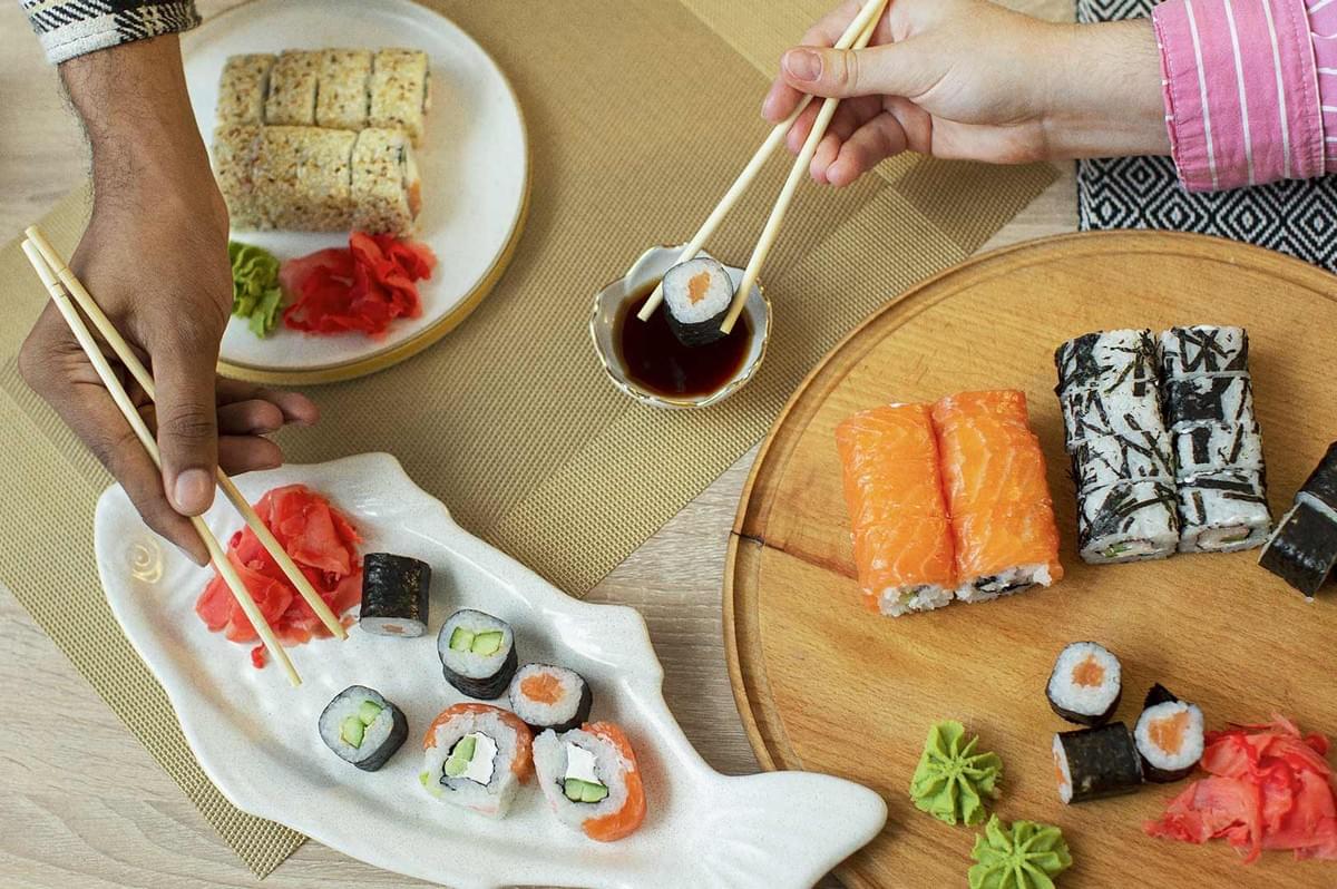 Assorted sushi platter with salmon-topped rolls, seaweed-wrapped maki, pickled ginger and wasabi; two hands using chopsticks to dip a roll into soy sauce on a shared dining table.