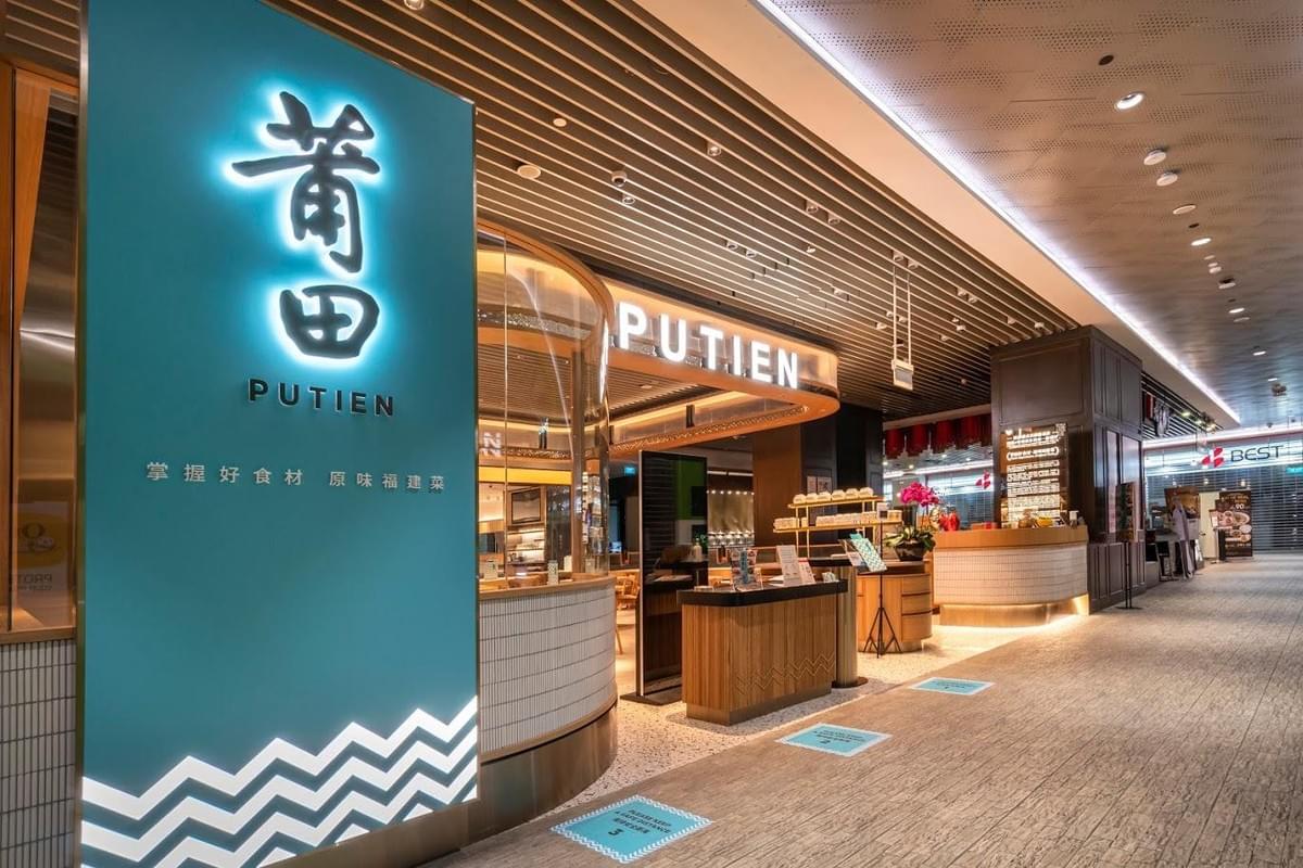 The image displays the modern storefront of a Putien restaurant located within a brightly lit shopping mall. A large, teal pillar features the restaurant's glowing signage in both Chinese characters and English text.