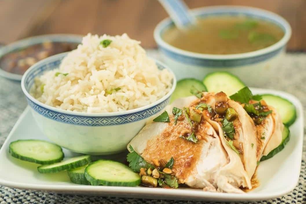 A steaming bowl of fragrant rice is served alongside tender chicken drizzled with a savory ginger-soy sauce and fresh cucumber slices. The meal is completed with a side of clear broth in a matching blue-and-white patterned bowl.