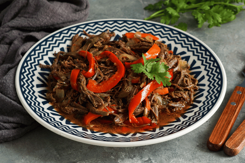 A wide shot of a decorative blue and white patterned plate holding a generous portion of shredded beef Ropa Vieja, mixed with sautéed onions and large, bright red pepper slices, garnished with cilantro.
