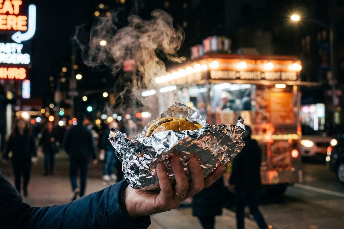 A hand holding a steaming cheeseburger wrapped in foil with a late-night food truck in the background.