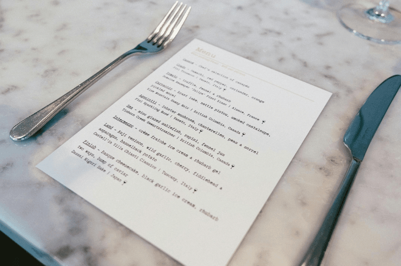 A fine-dining tasting menu printed on white paper resting on a marble table, positioned between a silver fork and knife, detailing upscale courses like miso-glazed sablefish and Koji venison.