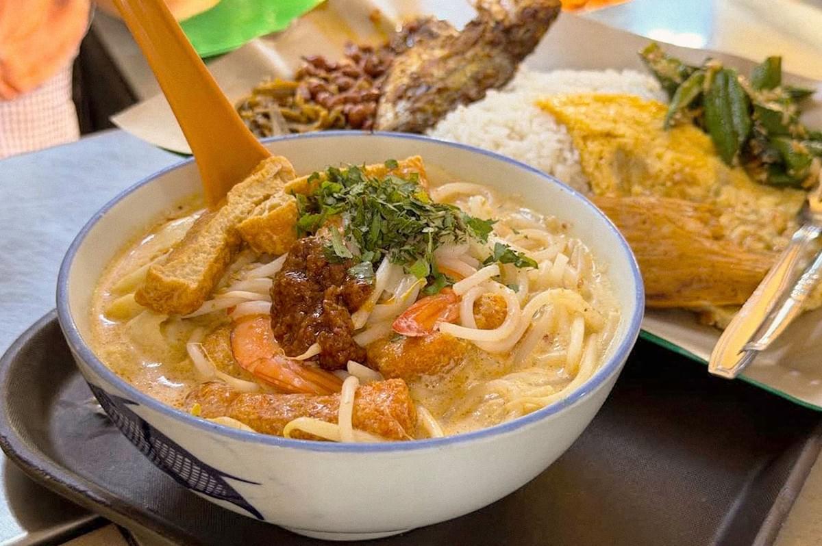 A bowl of creamy curry laksa topped with shrimp, tofu, fish cake, meatball, herbs, and rice noodles, served alongside a plate of rice and side dishes.