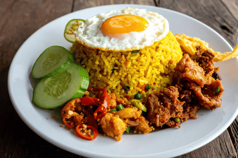 A top-down view of a hearty portion of turmeric-infused Nasi Goreng on a white plate, featuring a fried egg, fresh cucumber, and savory side portions of chili-fried chicken and vegetables.