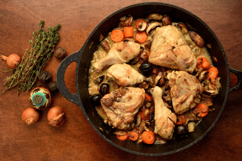 A top-down view of chicken pieces, sliced carrots, and mushrooms simmering in a light wine sauce in a black cast-iron pot, flanked by fresh thyme and shallots on a wooden surface.