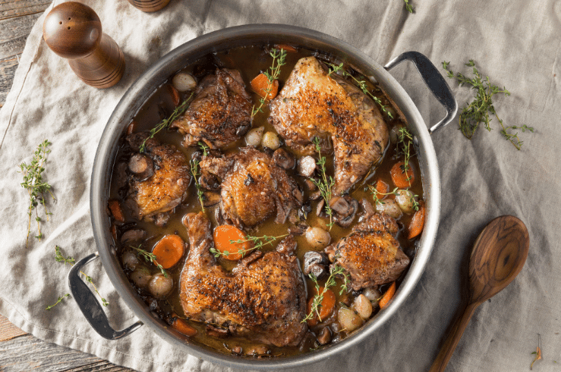 A large stainless steel pan of Coq au Vin with braised chicken thighs and legs garnished with fresh sprigs of thyme.