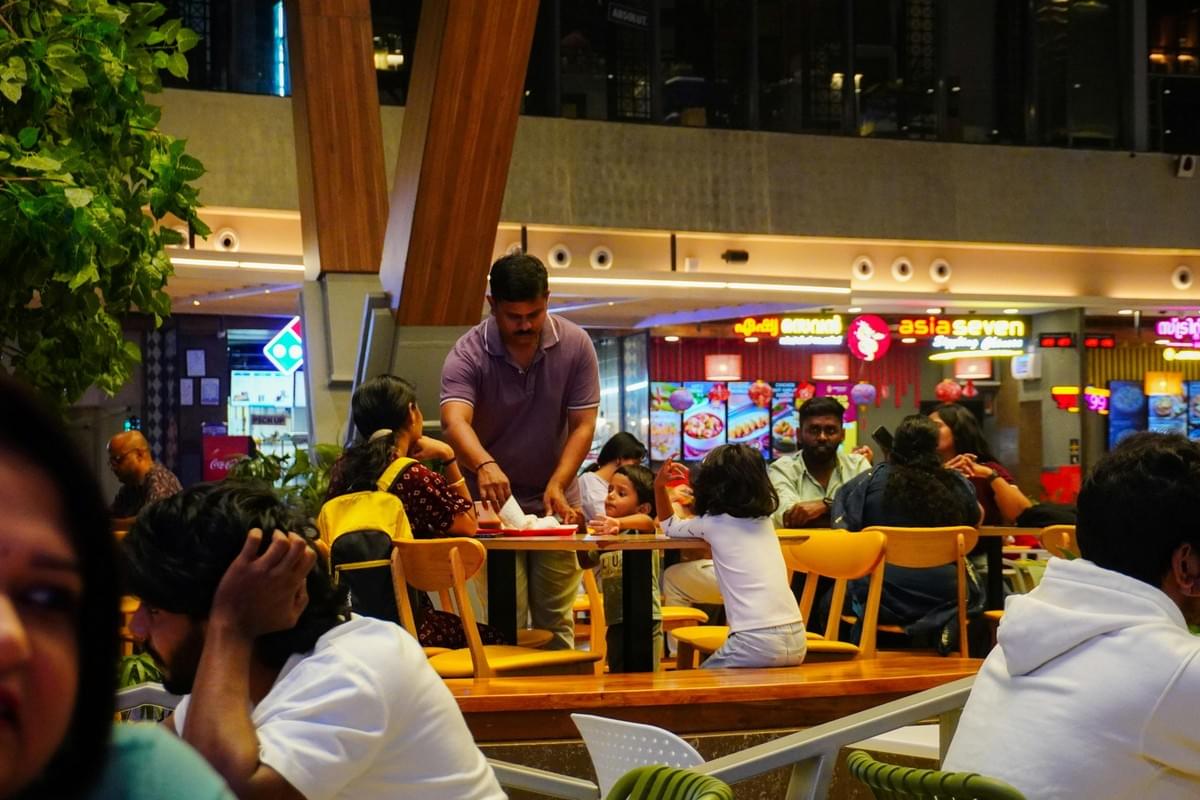 The image shows a bustling indoor food court or restaurant area where several people are seated at tables. In the center, a man in a purple polo shirt stands by a table with young children, while illuminated restaurant signs like "asia seven" are visible in the background.