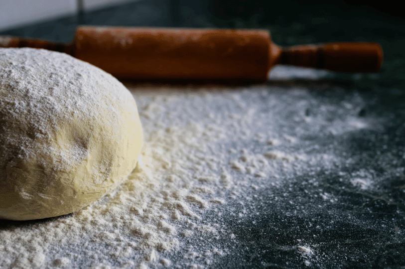 A smooth mound of proofing bread dough next to a wooden rolling pin, highlighting the elastic surface tension created by proper gluten development.