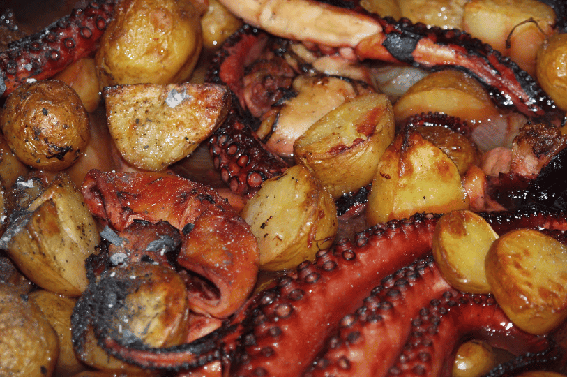 A close-up shot of a seafood Peka showing tender, charred octopus tentacles and golden-brown roasted potatoes glistening in savory cooking juices.