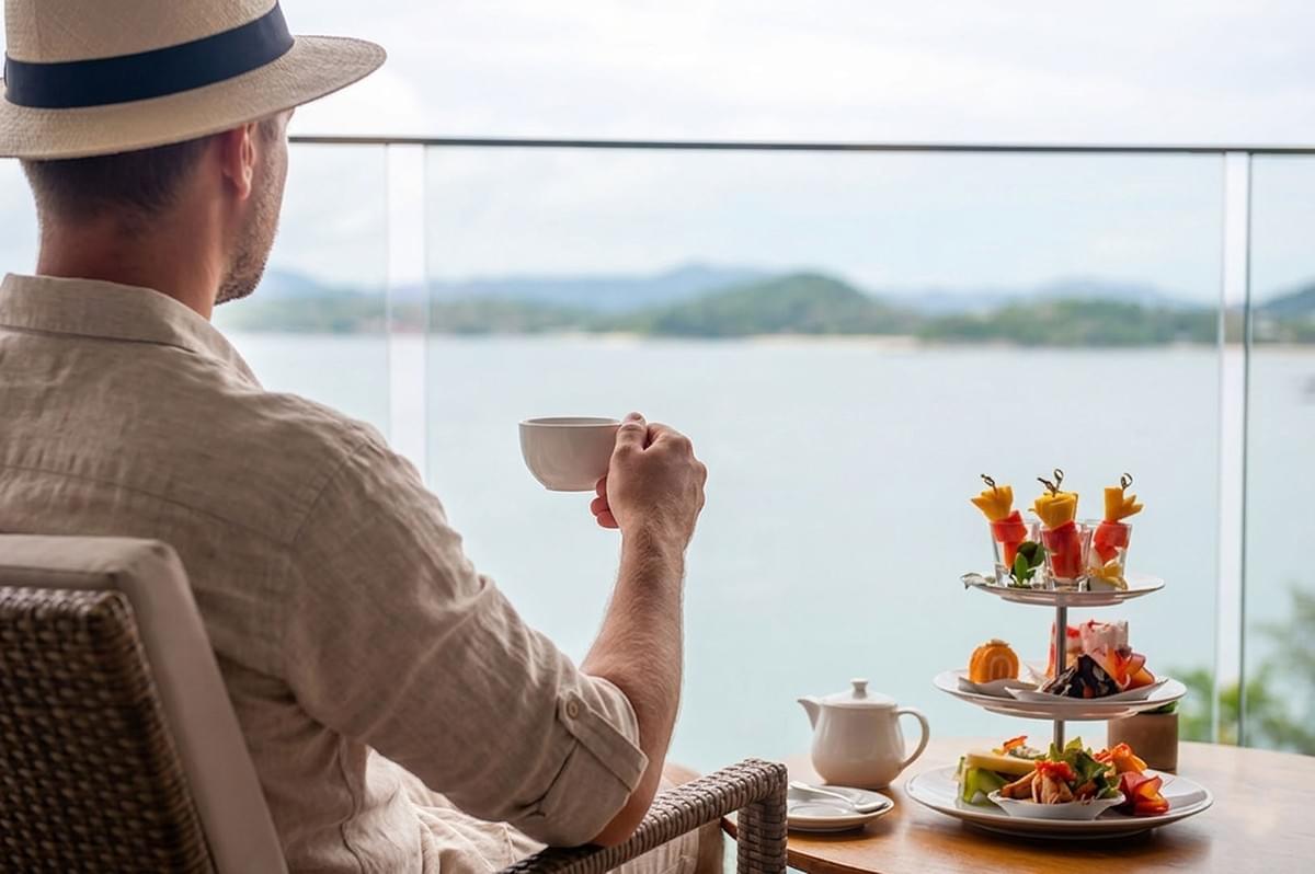 Man in straw hat enjoying afternoon tea with tiered pastry stand on balcony overlooking ocean and mountains