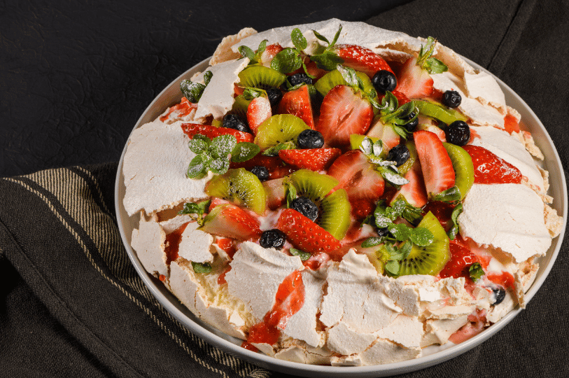 A rustic, large pavlova with a shattered, crispy meringue crust, piled high with sliced kiwi, strawberries, blueberries, and fresh mint.
