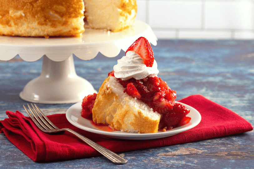 A slice of angel food cake topped with whipped cream and fresh strawberries sits on a white plate beside a red napkin and fork, with the remaining cake displayed on a pedestal in the background—capturing the dessert’s airy texture and vibrant, summery appeal.