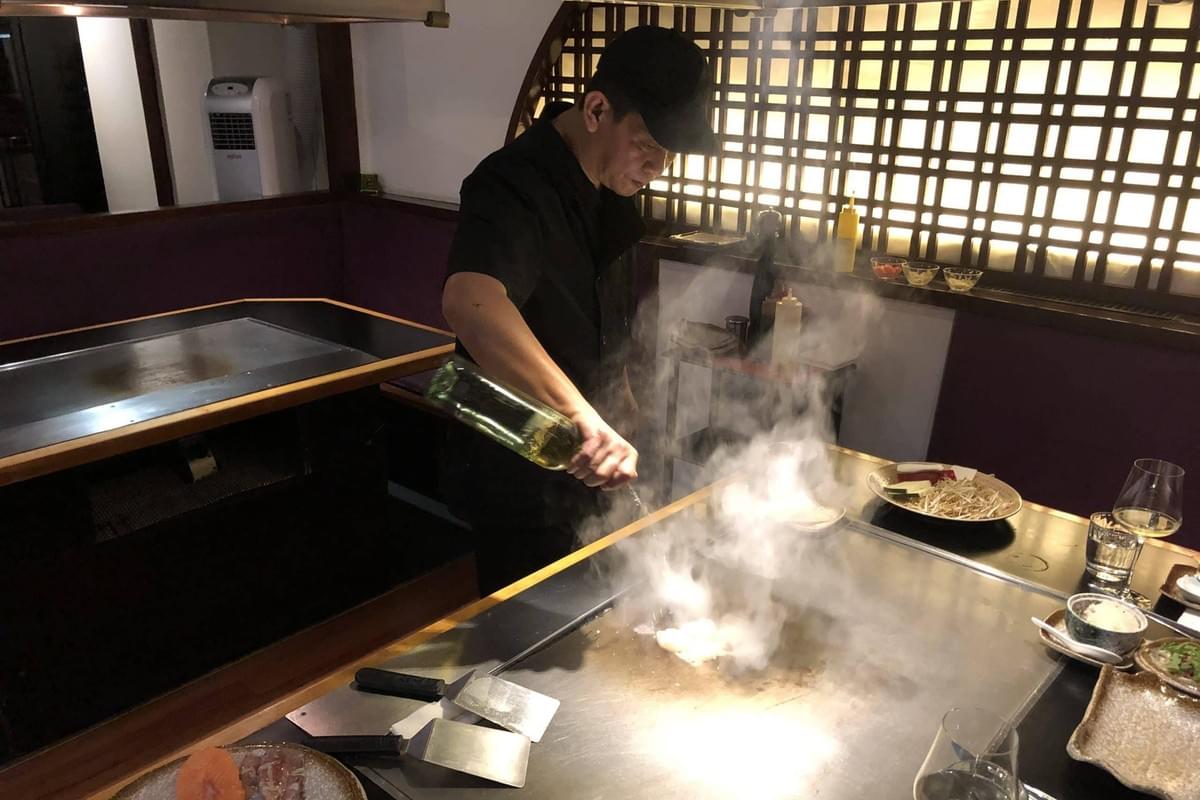 A chef wearing a black uniform and cap pours liquid onto a hot teppanyaki grill, creating a cloud of steam. Dishes of raw ingredients and wine glasses surround the flat-top stove in a dimly lit restaurant setting.