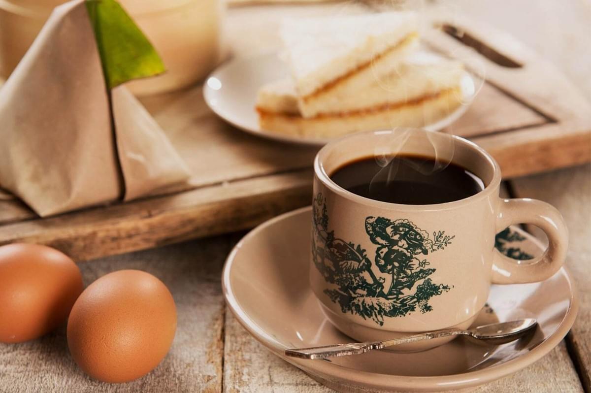 A steaming cup of coffee in a beige cup with green floral design is on a saucer beside a spoon; nearby are two boiled eggs and sandwich slices on a tray.