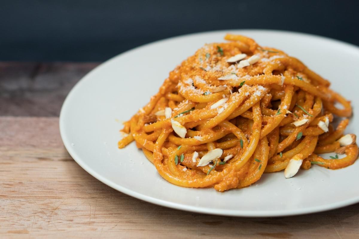 A plate of thick spaghetti-like pasta is coated in a vibrant orange sauce, garnished with slivered almonds, grated cheese, and fresh herbs. The dish is presented on a white plate resting on a wooden surface against a dark, blurred background.