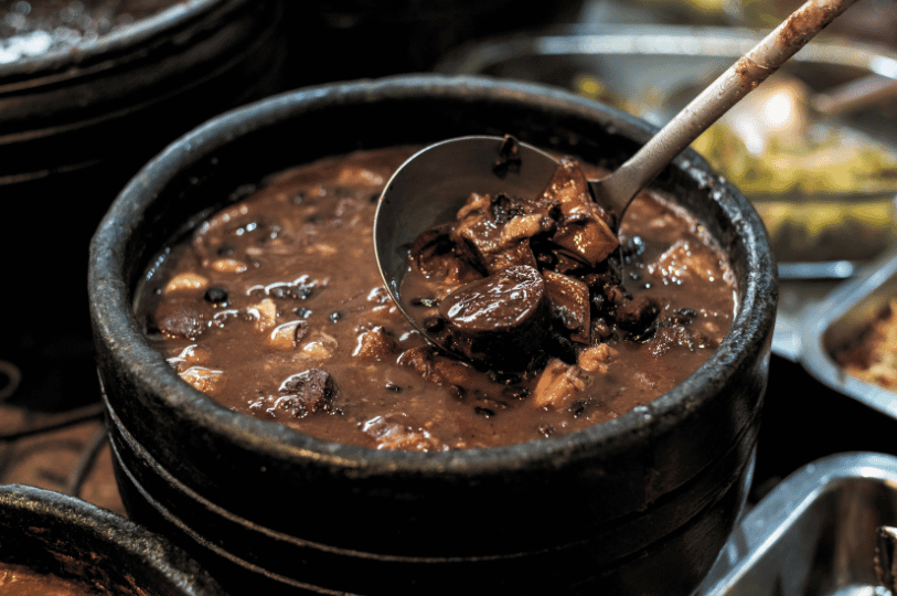 A black clay pot filled with rich Brazilian feijoada—black beans and assorted pork cuts—sits in a buffet-style setting, with a ladle revealing the stew’s hearty texture and celebratory spirit.