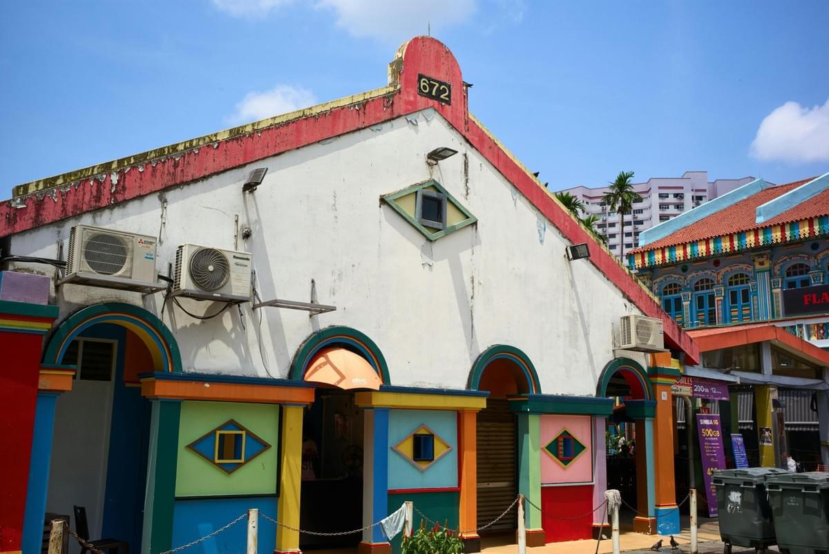 A vibrant, white-walled building features a distinctively shaped roofline marked with the number 672 and a small, diamond-shaped window resembling an eye. The building's lower level is adorned with a playful variety of bright colors, arched entryways, and geometric cutouts under a clear blue sky.