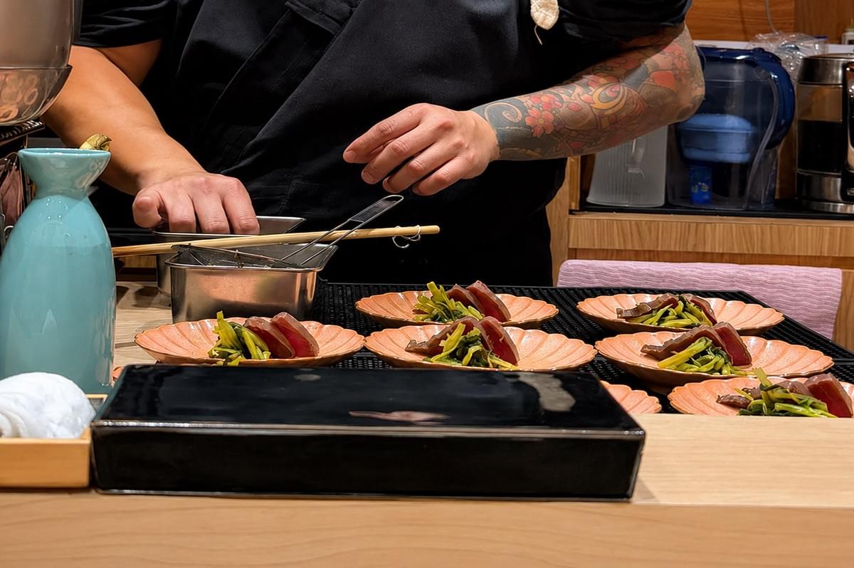 A chef wearing a black uniform and colorful floral sleeve tattoos stands behind a light-wood sushi counter, carefully plating dishes during service. In the middle ground, several peach-colored, flower-shaped ceramic plates are lined up on a black wire rack, each topped with a delicate portion of seared fish resting against vibrant cooked green vegetables. The foreground features a sleek, dark black rectangular tray alongside a rolled white hand towel in a wooden holder and a light blue ceramic sake carafe, while kitchen equipment like stainless steel pots and water pitchers are visible in the background background.