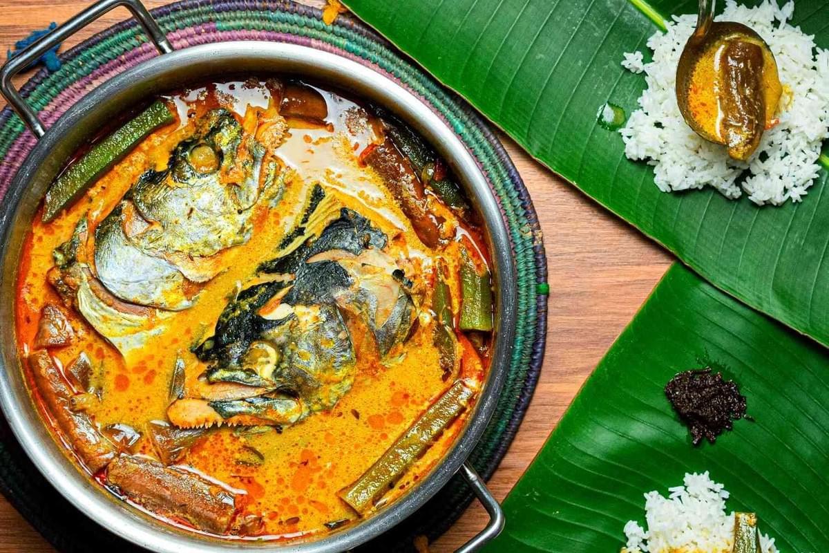 An overhead shot ofshows a large metal pot filled with a vibrant, orange fish head curry containing okra and eggplant. Next to the pot, portions of white rice and sambal are served on bright green banana leaves.