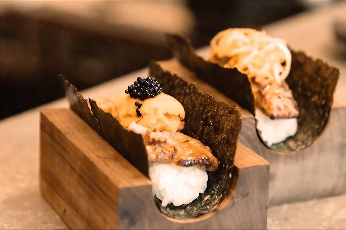 Two sushi tacos in wooden holders feature rice, crispy seaweed, topped with grilled fish, black caviar, and garnished with thinly sliced vegetables.