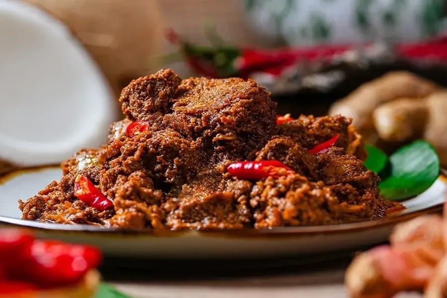 A plate of slow-cooked beef rendang is presented with a rich, dark spice paste and topped with fresh red chili slices. The dish is surrounded by its raw ingredients, including whole coconut, fresh ginger root, and kaffir lime leaves.