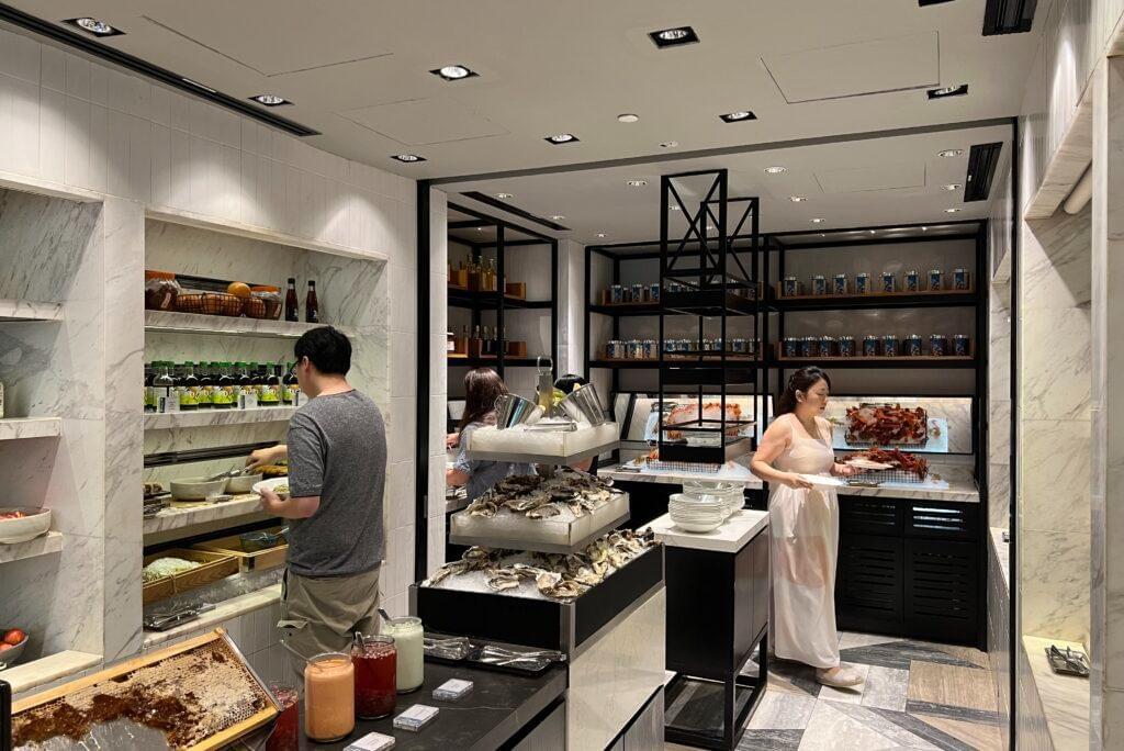 Guests browse food stations at a modern, upscale buffet featuring marble walls and black shelving. The display includes tiered fresh oysters on ice, bottled condiments, and a honeycomb station in the foreground.